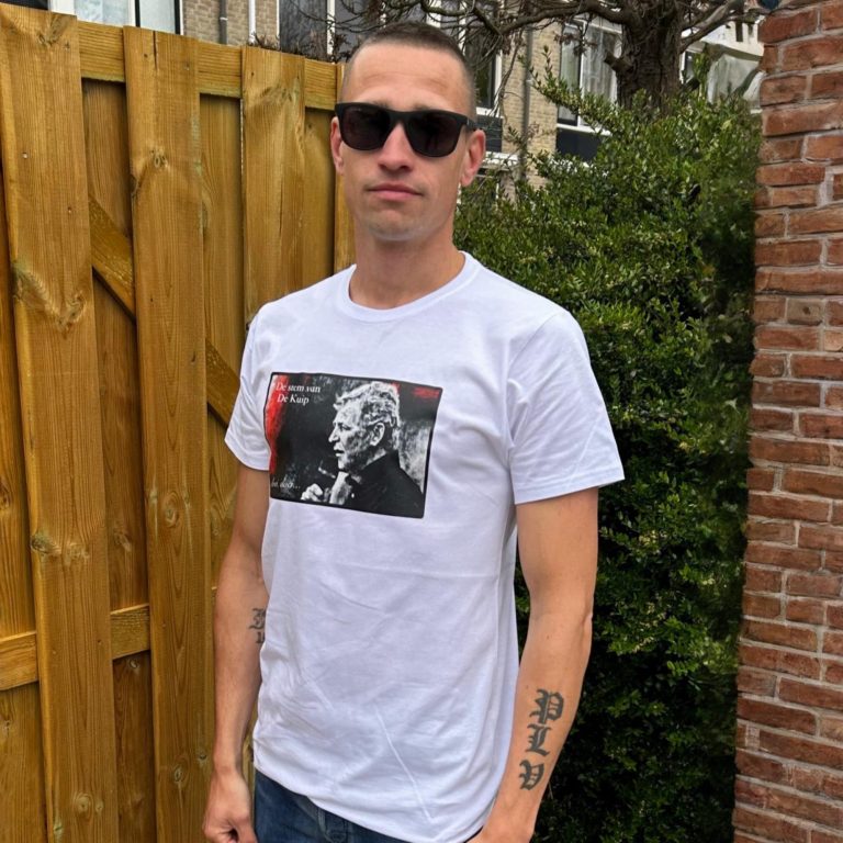 Man met een wit T-shirt en zonnebril, staand voor een houten hek.