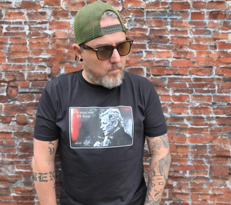 Man met een baard en tattoos, draagt een zwart T-shirt en een groene pet, staand tegen een bakstenen muur.
