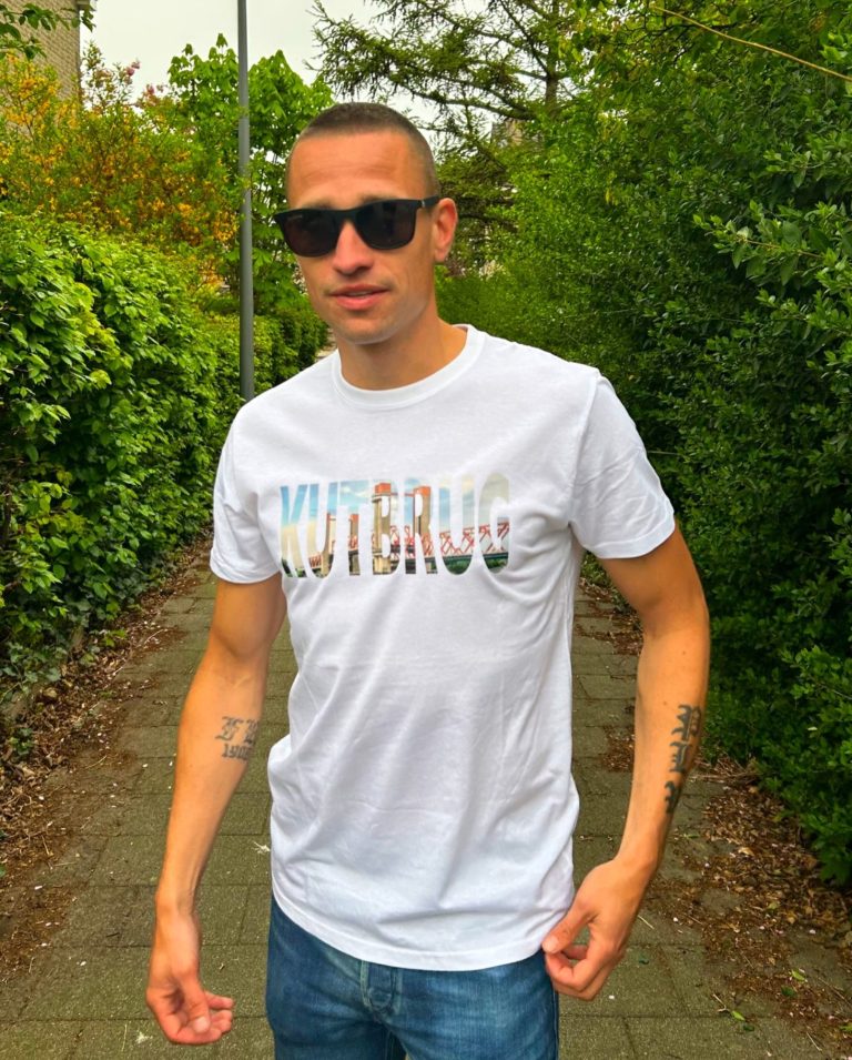 Jonge man in een wit t-shirt met de tekst "KUTBRUG", staand op een pad tussen bomen.