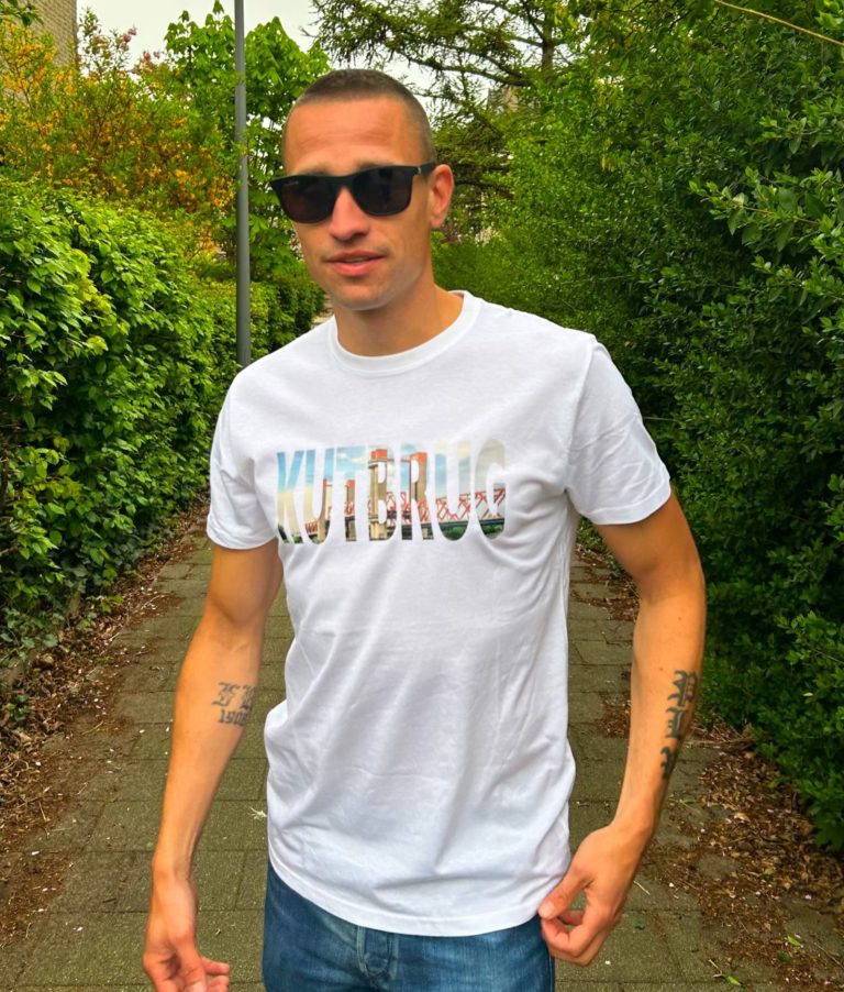 Jonge man in een wit T-shirt met kleurrijke tekst, buiten met groene achtergrond.