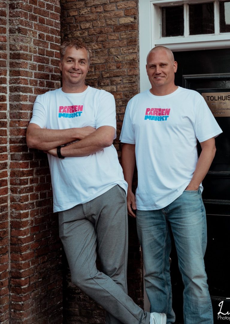 Twee mannen in witte T-shirts staan naast elkaar tegen een bakstenen muur.