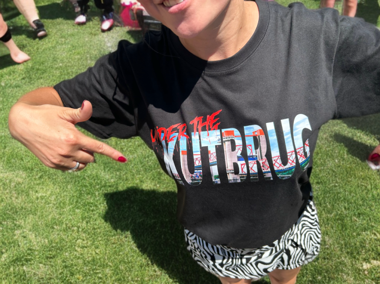 under the kutbrug t-shirt Vrouw in een zwart T-shirt met de tekst "under the kustbrug" wijst ernaar.