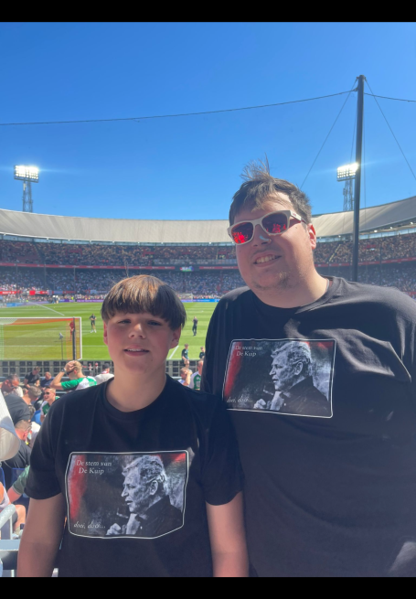 Twee mensen staan op een stadion, beiden dragen een zwart shirt met een afbeelding.