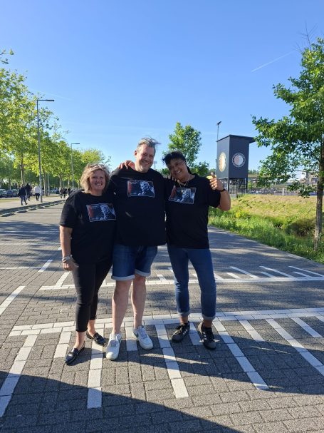 Drie mensen in zwarte T-shirts poseren lachend op een parkeerplaats.