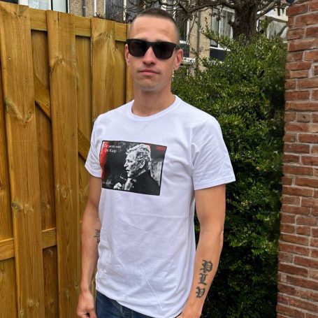 Jonge man in witte T-shirt met afbeelding, zonnebril en tatoeage, voor houten schutting.