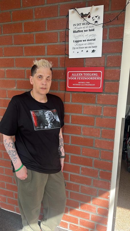 Persoon met kort, blond haar en tattoo's staat voor een bakstenen muur met borden.