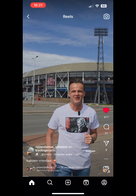 Man in een wit t-shirt poseert voor een stadion onder een blauwe lucht.