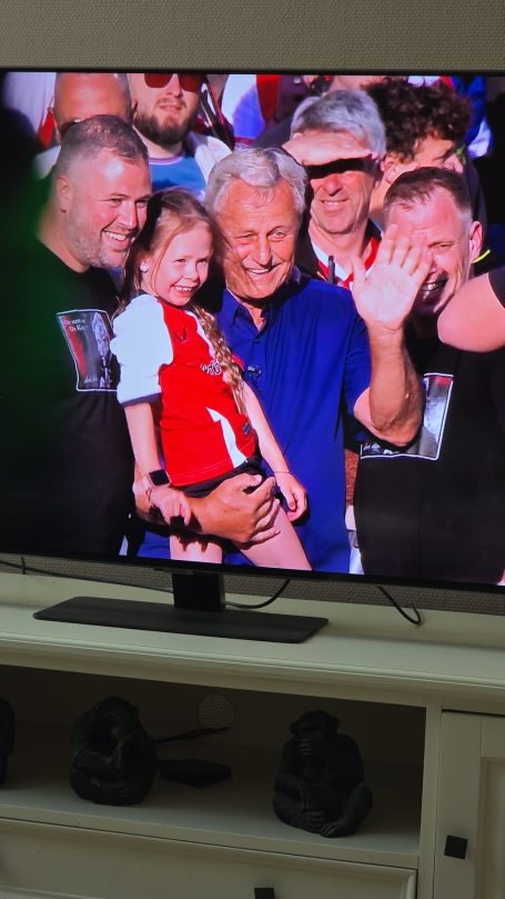 Groep mensen, waaronder een man met grijs haar en een jong meisje, die glimlachen en zwaaien.