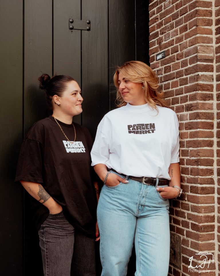 Twee vrouwen staan naast elkaar, gekleed in T-shirts en spijkerbroeken, met een bakstenen muur op de achtergrond.