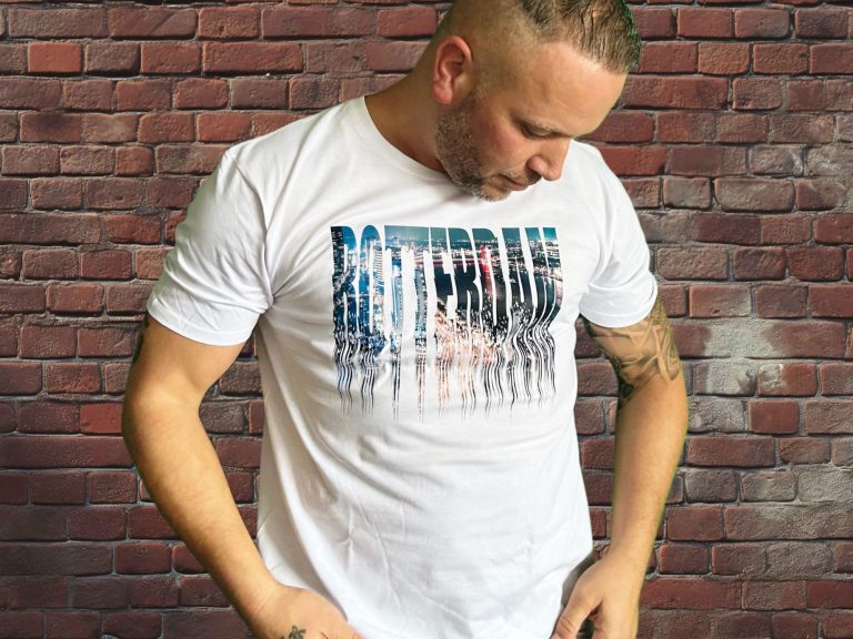 Man in wit T-shirt met kleurrijke opdruk, staande voor een bakstenen muur.