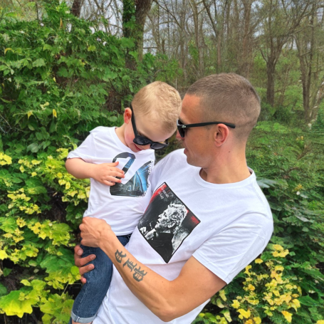 Vader en zoon in witte T-shirts, samen met zonnebrillen, omringd door groene bladeren.